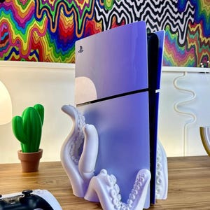 May include: A white octopus-shaped stand holding a purple PlayStation console. The console is standing vertically and has the PlayStation logo on the front. The stand is on a wooden surface.