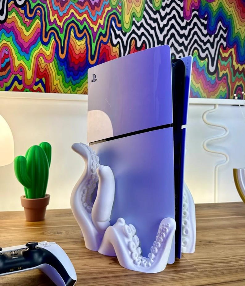 May include: A white octopus-shaped stand holding a purple PlayStation console. The console is standing vertically and has the PlayStation logo on the front. The stand is on a wooden surface.