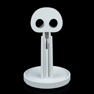 May include: A white, stylized skull-shaped object with two large eye sockets and a vertical stem. The stem is connected to a circular base with a ridged edge. The object is made of a solid, opaque material and is set against a black background.