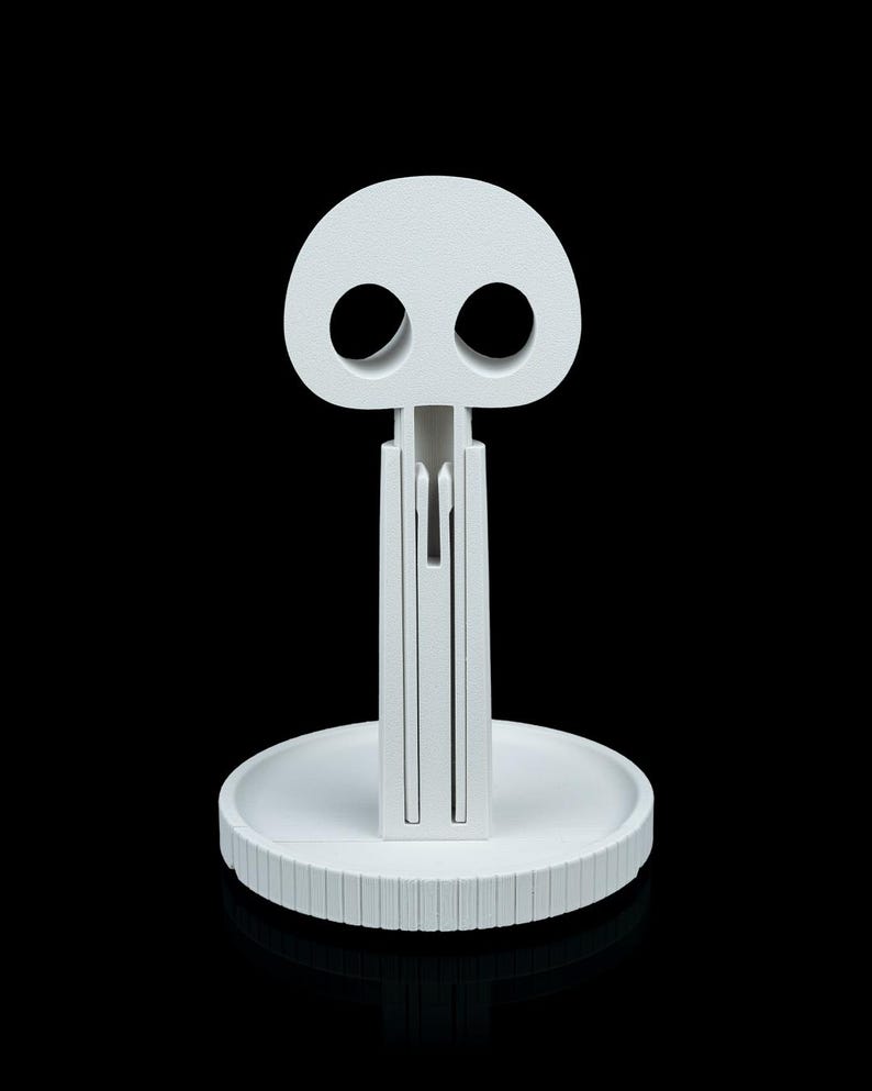 May include: A white, stylized skull-shaped object with two large eye sockets and a vertical stem. The stem is connected to a circular base with a ridged edge. The object is made of a solid, opaque material and is set against a black background.