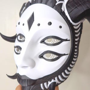 May include: A white and black mask with a stylized face, featuring multiple eyes and a diamond-shaped detail. The mask has curved horns and intricate detailing around the edges, creating a striking and artistic design.