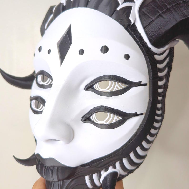 May include: A white and black mask with a stylized face, featuring multiple eyes and a diamond-shaped detail. The mask has curved horns and intricate detailing around the edges, creating a striking and artistic design.