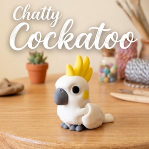 May include: A small, white cockatoo figurine with a bright yellow crest, a grey beak, and yellow cheek patches. The bird has black eyes and grey feet. The text "Chatty Cockatoo" is displayed in white script above the figurine on a light background.