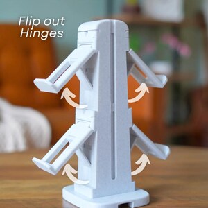 May include: A light gray plastic object with flip-out hinges. The object has a rectangular base and a central column with multiple hinged arms extending outwards. The text "Flip out Hinges" is visible in the upper left corner. The object is on a wooden surface.