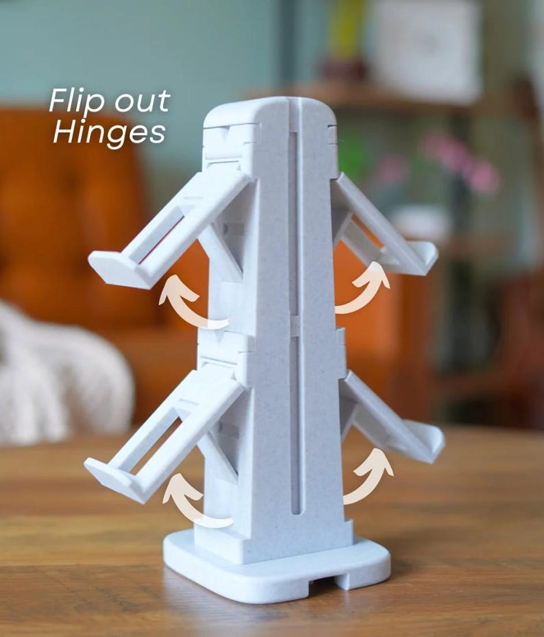 May include: A light gray plastic object with flip-out hinges. The object has a rectangular base and a central column with multiple hinged arms extending outwards. The text "Flip out Hinges" is visible in the upper left corner. The object is on a wooden surface.