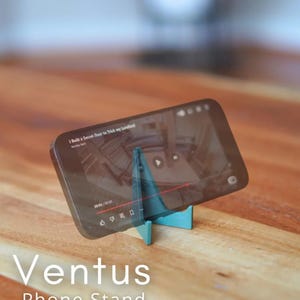 Może przedstawiać: Turkusowy stojak na telefon podtrzymujący smartfon z filmem. Stojak stoi na drewnianej powierzchni. Tekst "Ventus Phone Stand" jest widoczny poniżej. Film pokazuje sekretne drzwi.
