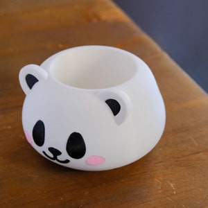 May include: White ceramic planter shaped like a panda bear with black eyes, nose, and ears. The planter has pink cheeks.