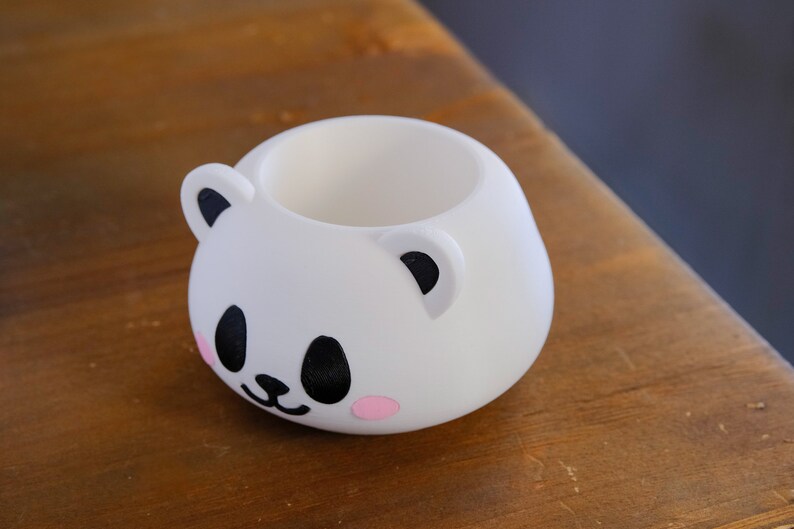 May include: White ceramic planter shaped like a panda bear with black eyes, nose, and ears. The planter has pink cheeks.