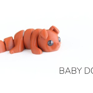 May include: A small, brown, segmented, articulated toy dog with black eyes. The toy is lying on its side with its head turned to the right. The toy is labelled "BABY DOG".