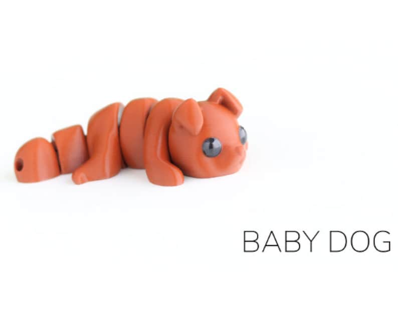 May include: A small, brown, segmented, articulated toy dog with black eyes. The toy is lying on its side with its head turned to the right. The toy is labelled "BABY DOG".