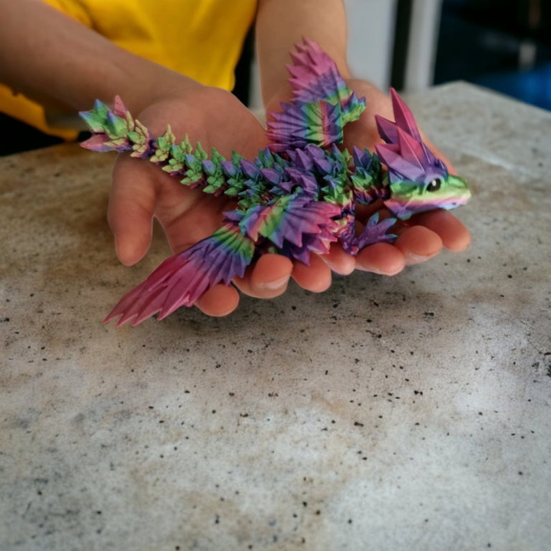 K&ouml;nnte beinhalten: Ein bunter Origami-Drache aus Papier, mit rosa, lila, gr&uuml;nen und blauen Akzenten. Der Drache wird in den H&auml;nden einer Person gehalten.