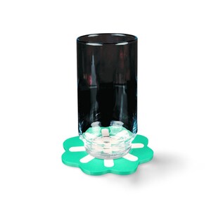 May include: A tall, black glass sits atop a green, flower-shaped coaster with white accents. A small, clear glass object rests on the coaster. The background is plain white.
