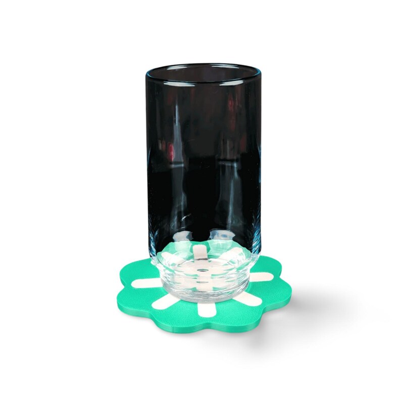 May include: A tall, black glass sits atop a green, flower-shaped coaster with white accents. A small, clear glass object rests on the coaster. The background is plain white.