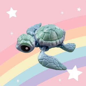 Puede incluir: Una figurita de tortuga impresa en 3D de color azul claro y verde con una concha detallada y un ojo negro. La tortuga est&aacute; posada sobre su vientre con las patas y la cabeza extendidas.