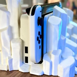 May include: A white 3D printed stand with a white Nintendo Switch console and controller. The stand is designed to look like a rocky mountain range.