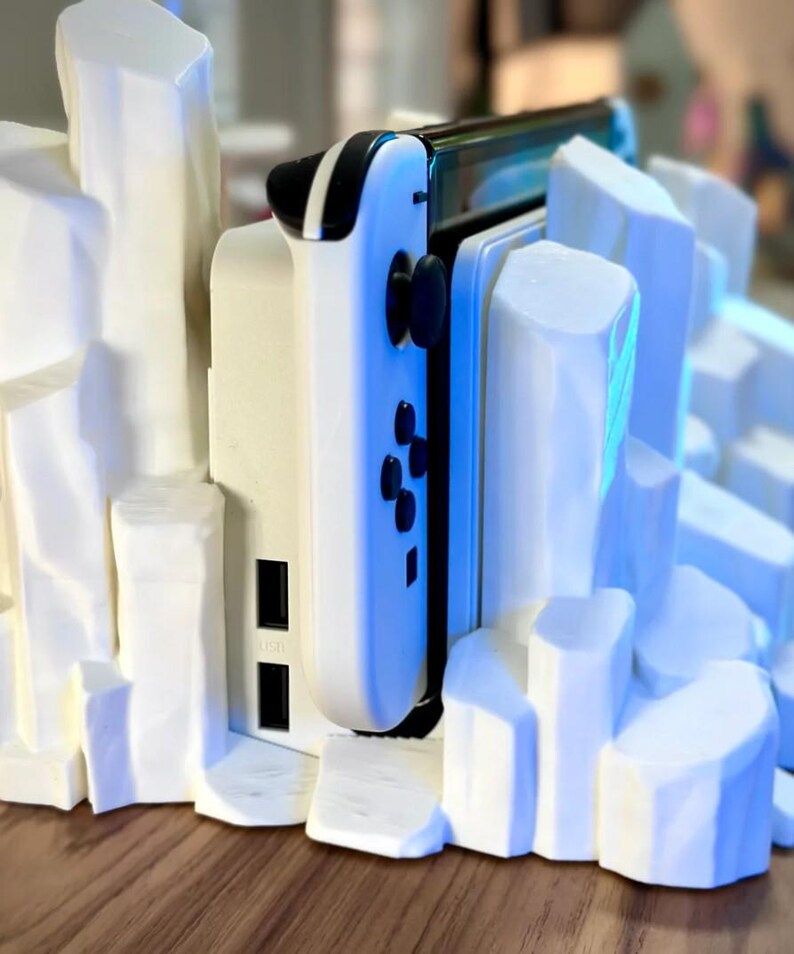 May include: A white 3D printed stand with a white Nintendo Switch console and controller. The stand is designed to look like a rocky mountain range.