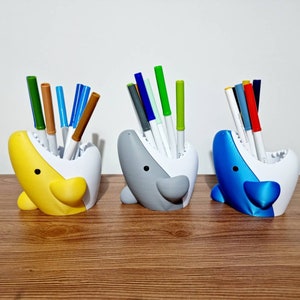 May include: Three colorful shark-shaped desk organizers with open mouths, each holding a variety of colored pens and markers.
