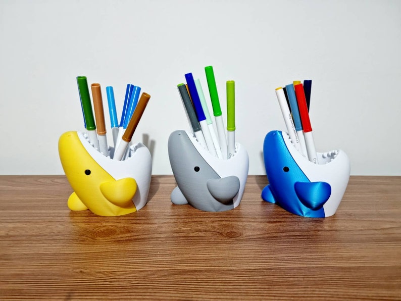 May include: Three colorful shark-shaped desk organizers with open mouths, each holding a variety of colored pens and markers.