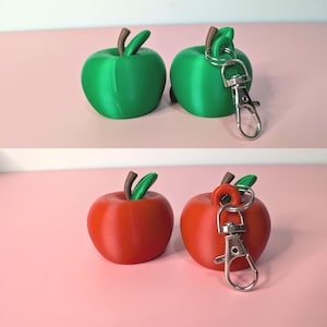 May include: Green and red apple-shaped keychains. The green apples have brown stems and green leaves. The red apples have brown stems and green leaves. Each keychain has a silver-colored clasp.