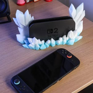 May include: A black Nintendo Switch dock with a decorative, icy-crystal-shaped stand in white and light blue. A black Nintendo Switch console is in the foreground, with orange and blue accents.