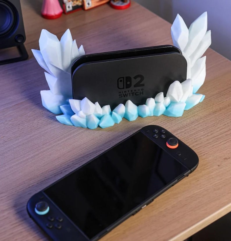 May include: A black Nintendo Switch dock with a decorative, icy-crystal-shaped stand in white and light blue. A black Nintendo Switch console is in the foreground, with orange and blue accents.