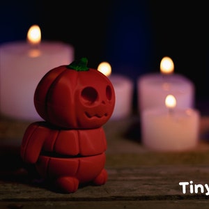 May include: A small, red, pumpkin-shaped figurine with a carved jack-o'-lantern face. The figurine is standing on a wooden surface in front of several lit candles.