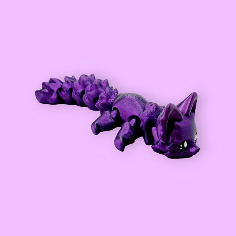 Puede incluir: Una figurita de zorro articulada impresa en 3D de color morado con una cola tupida. El zorro est&aacute; acostado boca abajo con la cabeza girada hacia un lado.
