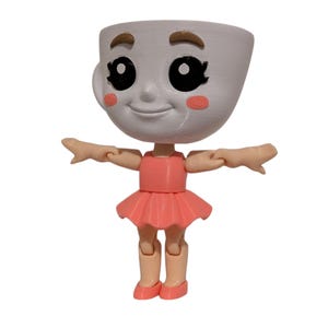 May include: A whimsical figurine of a teacup with a smiling face, rosy cheeks, and large black eyes. The teacup head is attached to a body wearing a pink dress and ballet shoes. The arms are outstretched.