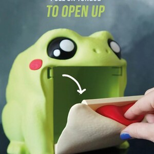 May include: A light green frog-shaped container with large black and white eyes and red cheek spots. The text "PULL ON TONGUE TO OPEN UP" is above the frog. A hand pulls the red tongue to open the container.