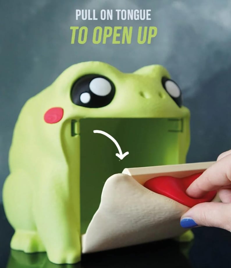 May include: A light green frog-shaped container with large black and white eyes and red cheek spots. The text "PULL ON TONGUE TO OPEN UP" is above the frog. A hand pulls the red tongue to open the container.