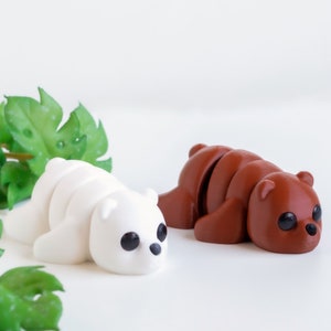 Puede incluir: Dos peque&ntilde;os juguetes de peluche de oso segmentados. Un oso es blanco y el otro es marr&oacute;n. Ambos osos est&aacute;n tumbados sobre una superficie blanca con hojas verdes en el fondo.