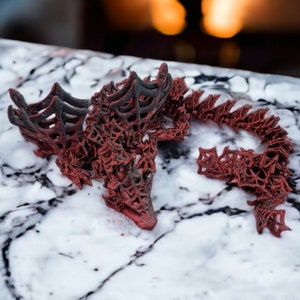 May include: A detailed red and black dragon sculpture with an openwork design. The dragon is positioned on a marble surface, with its head and tail in view. The figurine has a textured finish, highlighting the dragon's scales and wings.