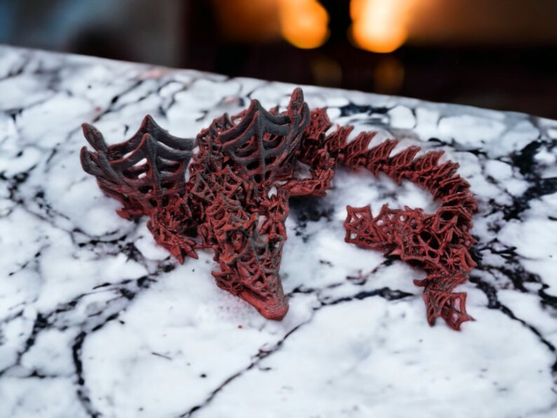 May include: A detailed red and black dragon sculpture with an openwork design. The dragon is positioned on a marble surface, with its head and tail in view. The figurine has a textured finish, highlighting the dragon's scales and wings.