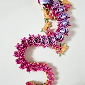 Peut inclure: Un dragon en tissu fait de roses, de couleur rose et violette. Le dragon est enroul&eacute; en forme de S, avec la t&ecirc;te en haut de l'image. Le dragon est compos&eacute; de nombreuses petites roses, et la queue est enroul&eacute;e.