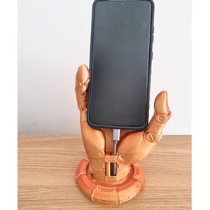 May include: A 3D printed phone stand in the shape of a robotic hand holding a smartphone. The hand is a metallic gold color and the phone is black.