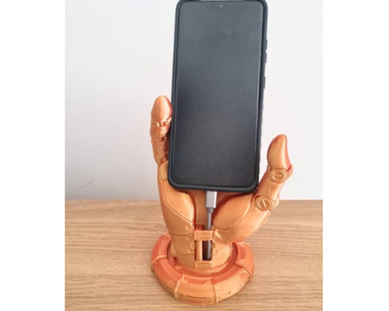 May include: A 3D printed phone stand in the shape of a robotic hand holding a smartphone. The hand is a metallic gold color and the phone is black.