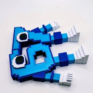 May include: A pixelated, blue and white 3D-printed figure with a square head and rectangular limbs. The figure has dark eyes and white hands. The figure is lying on a white surface.