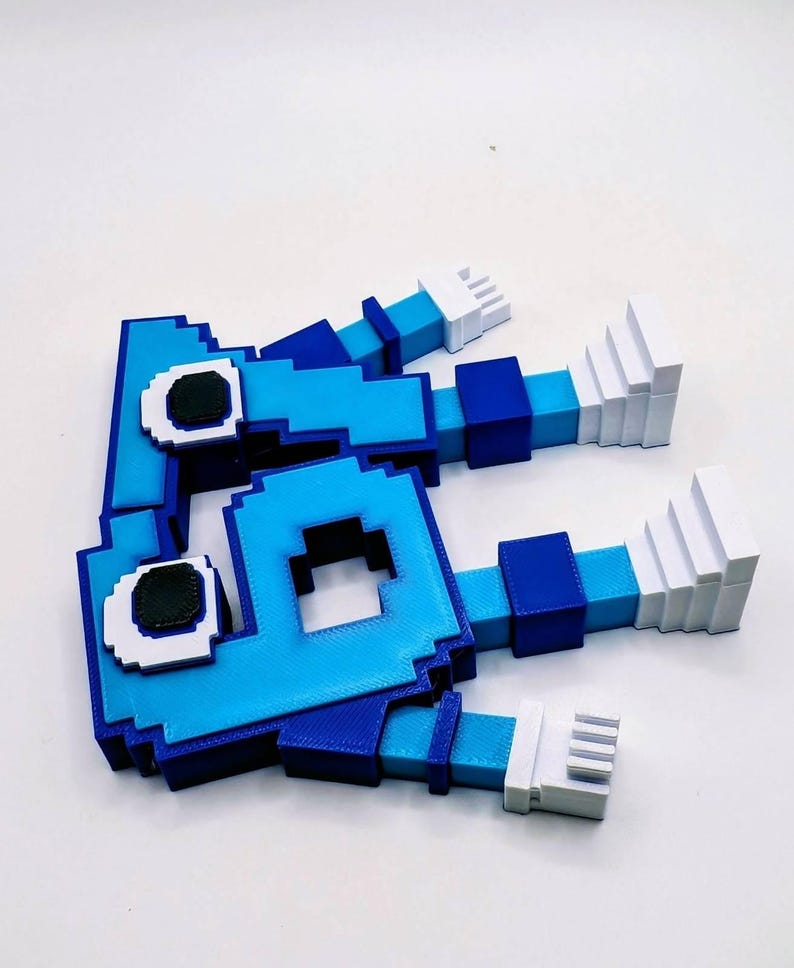 May include: A pixelated, blue and white 3D-printed figure with a square head and rectangular limbs. The figure has dark eyes and white hands. The figure is lying on a white surface.