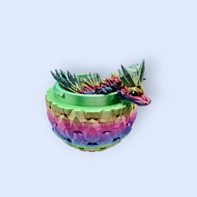 May include: A rainbow-colored dragon figurine is nestled inside a green and rainbow-colored pot. The dragon has a spiky back and a long tail.