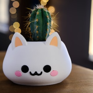 May include: White ceramic planter shaped like a cat's face with pink cheeks and black eyes, holding a small green cactus.