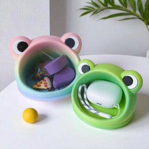 May include: Two frog-shaped containers, one rainbow-colored and the other green, sit on a white surface. The rainbow container holds purple sponges and a pizza slice. The green container holds a white earbud case. A yellow lemon is in the foreground.