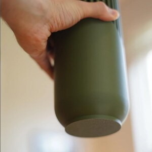 May include: An olive green cylindrical vase with vertical ribbed detailing near the top. The vase is held by a hand, showcasing its smooth, matte finish. The base is a lighter, textured color. A modern, minimalist design.