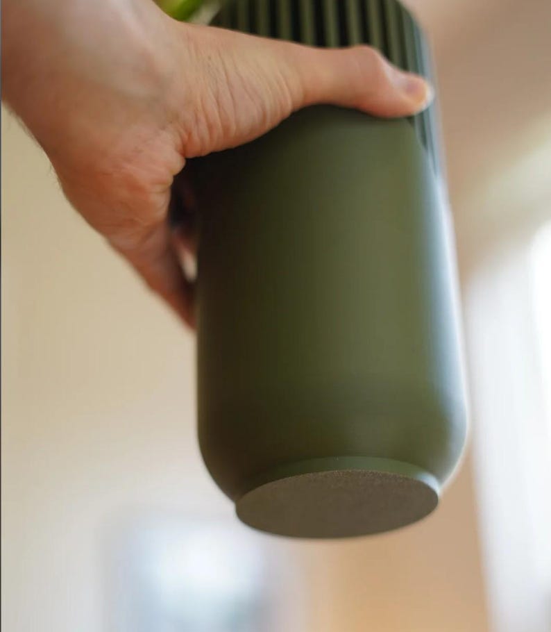 May include: An olive green cylindrical vase with vertical ribbed detailing near the top. The vase is held by a hand, showcasing its smooth, matte finish. The base is a lighter, textured color. A modern, minimalist design.