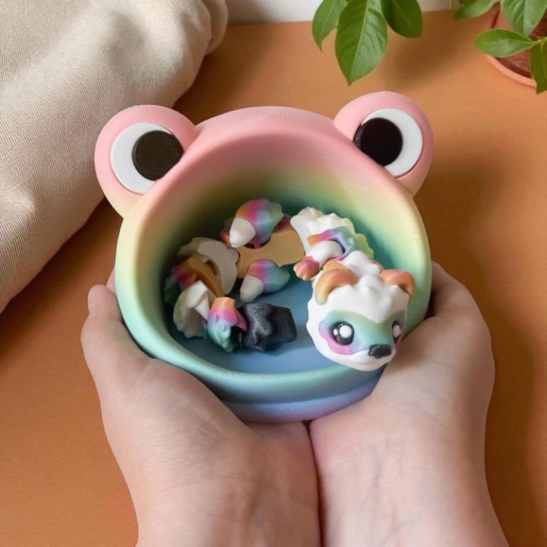 May include: A frog-shaped trinket dish in pastel colors of pink, yellow, and blue, filled with small, colorful, decorative items. The dish has large, round eyes and is held in two hands.