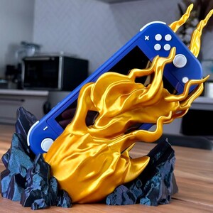 May include: A 3D printed stand for a Nintendo Switch Lite console, shaped like a hand emerging from a rocky base with flames. The console is blue and the flames are gold.