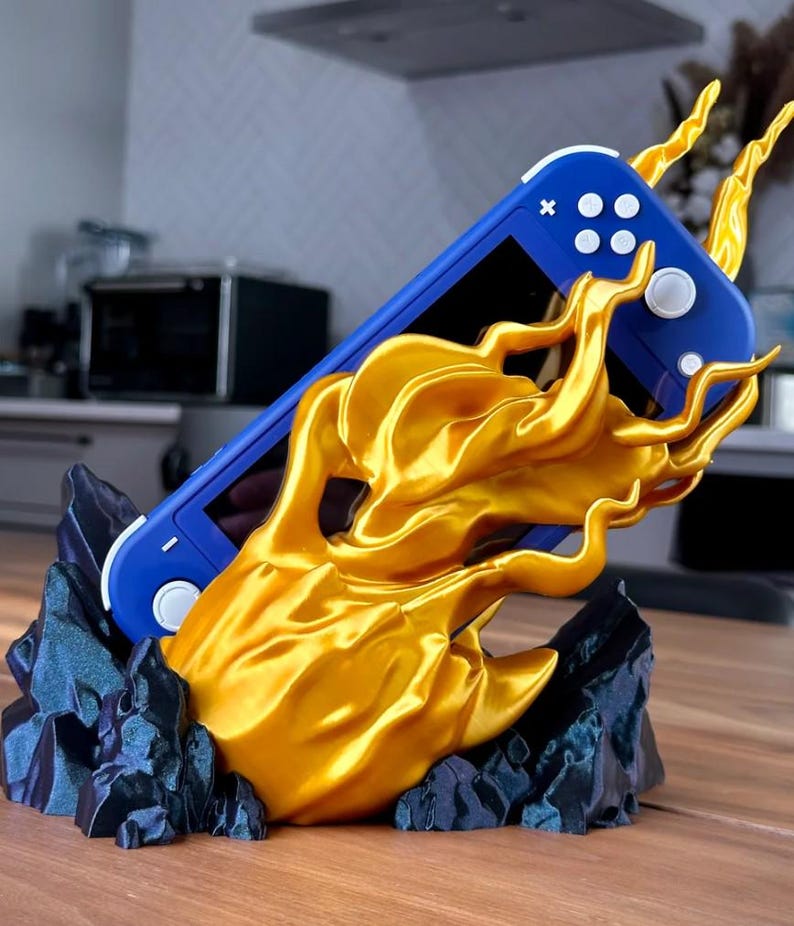 May include: A 3D printed stand for a Nintendo Switch Lite console, shaped like a hand emerging from a rocky base with flames. The console is blue and the flames are gold.