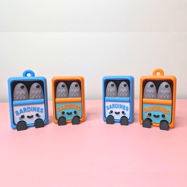 May include: Four whimsical sardine can figurines. Each can is a different color, including blue and orange, with a smiling face and the word "Sardines" on the front. The cans contain two gray fish.