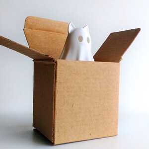 K&ouml;nnte beinhalten: Eine wei&szlig;e Keramik-Geisterfigur mit Katzenohren und ovalen Augenl&ouml;chern blickt aus einem braunen Pappkarton. Die oberen Klappen der Box sind ge&ouml;ffnet. Der Hintergrund ist schlicht wei&szlig;, was eine einfache, saubere &Auml;sthetik schafft.