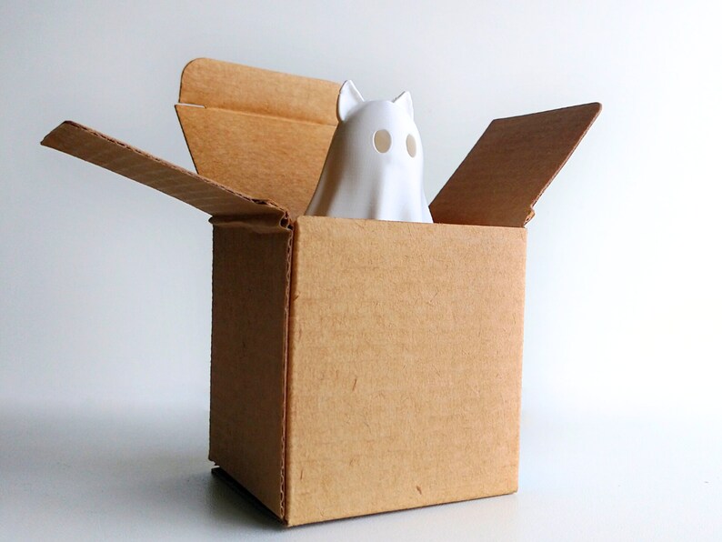 K&ouml;nnte beinhalten: Eine wei&szlig;e Keramik-Geisterfigur mit Katzenohren und ovalen Augenl&ouml;chern blickt aus einem braunen Pappkarton. Die oberen Klappen der Box sind ge&ouml;ffnet. Der Hintergrund ist schlicht wei&szlig;, was eine einfache, saubere &Auml;sthetik schafft.