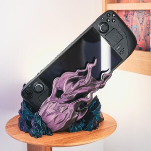 May include: A black handheld gaming device with a purple and blue 3D printed stand shaped like flames. The stand is on a wooden base.